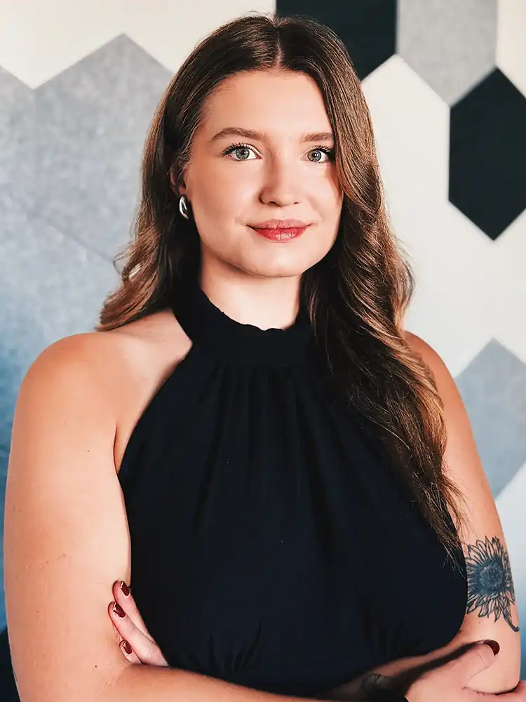 Woman with long brown hair wearing a black halter top, arms crossed, standing against a geometric patterned wall.
