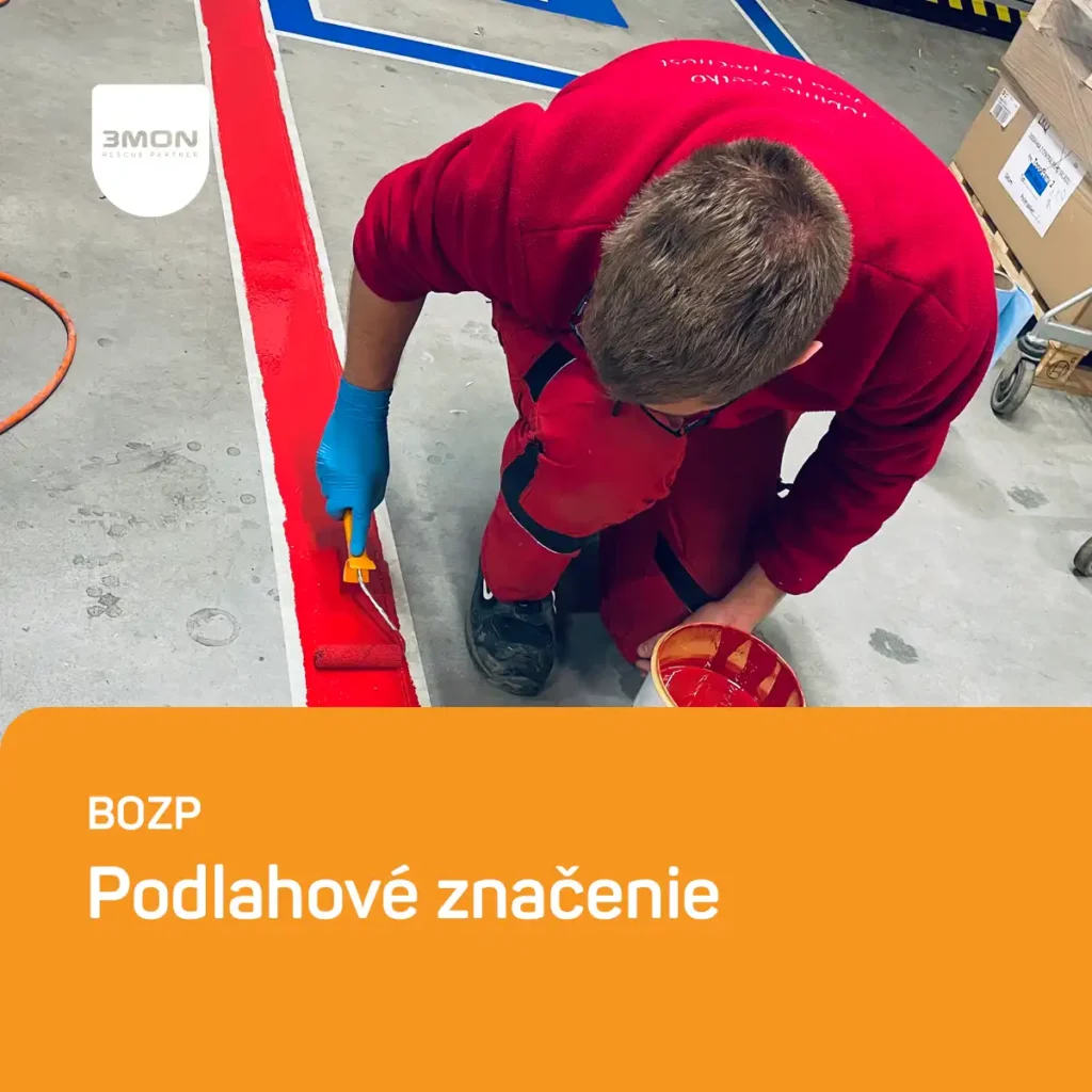 Worker in red overalls painting a bright red floor marking with a small roller brush and orange paint bucket in a workshop setting. Podlahové značenie
