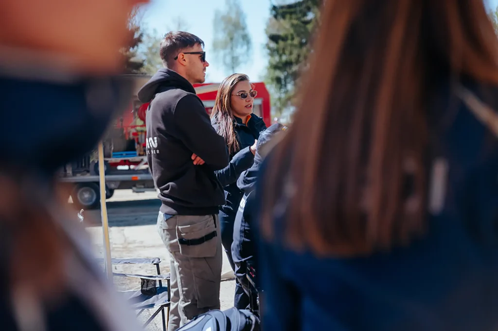 Two people wearing sunglasses and jackets talk outside at an event, framed by blurred onlookers in the foreground.