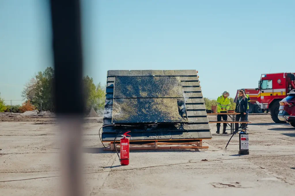Firefighters and a worker inspect a tilted solar panel array on a construction site, with a red fire extinguisher nearby and a fire truck in the background.