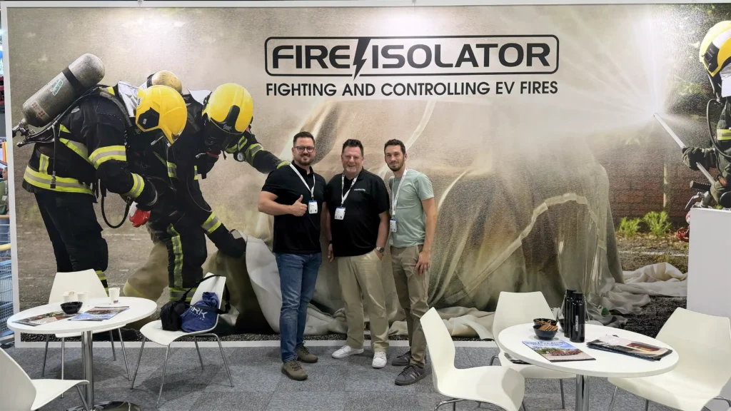 Three men with lanyards pose in front of a FireIsolator trade show backdrop featuring firefighters and the slogan 'FIRE ISOLATOR'.