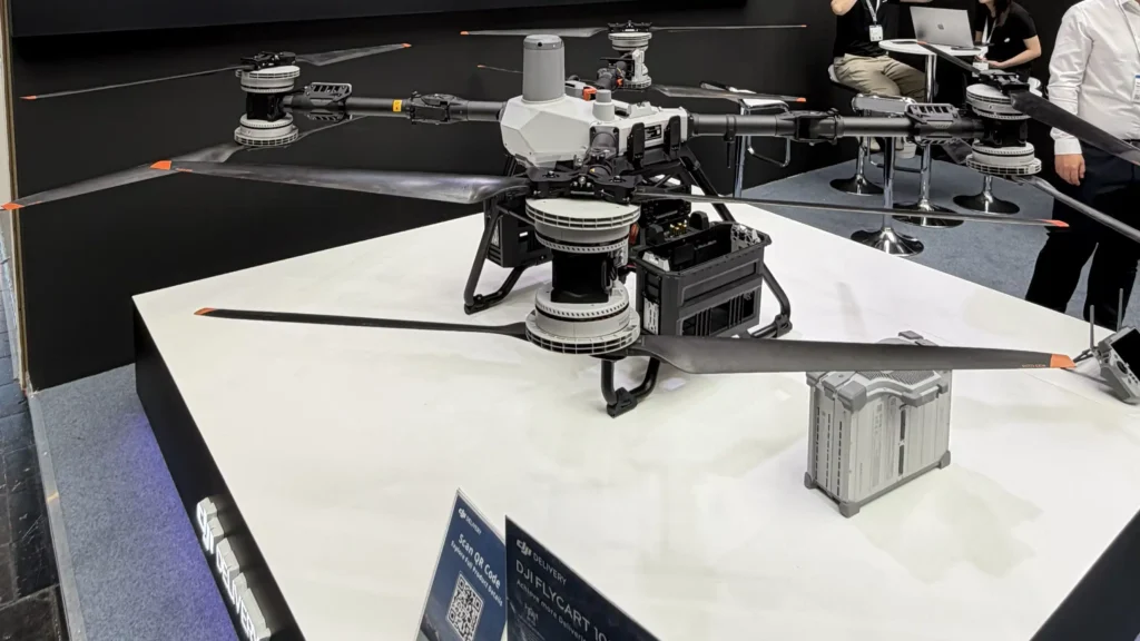 Large hexacopter drone on a white display table at a tech expo, with people and laptops in the background