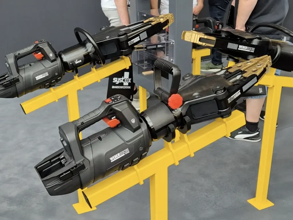 Three black industrial grippers mounted on a yellow stand in a workshop, with people visible in the background.